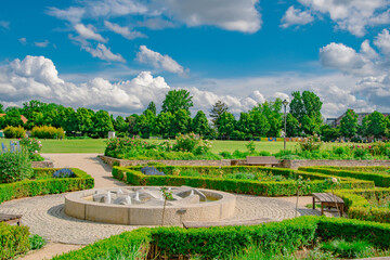Fototapeta premium Lush green cemetery garden in Ingolstadt, Bavaria, with blooming flowers, tombstones, and trees under a bright blue sky 