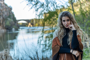 Portrait of a confident young woman with long wavy hair, wearing warm autumn clothes, standing by a...
