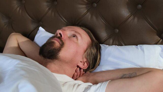 Young handsome Caucasian blond man with beard and mustache lying in bed, arms behind head, looking up at the ceiling with a thoughtful expression. Relaxed evening moment before sleep