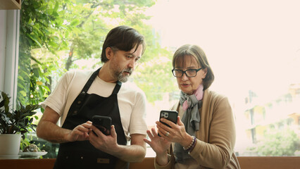 A friendly middle aged barista and a senior businesswoman collaborate using a smartphone and laptop...