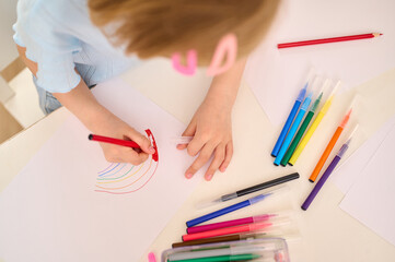 child drawing colorful rainbow with markers on white paper, expressing creativity. top view of...