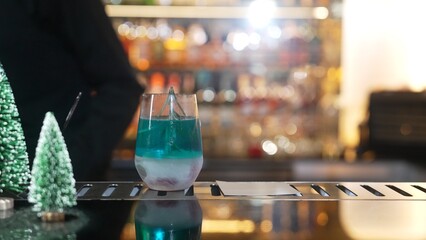 Bartender serving Christmas blue Lagoon cocktail with red dried fruit rosemary design on luxury bar counter with VIP group guest friend or couple in green neon snow Christmas tree festive. Vinosity.
