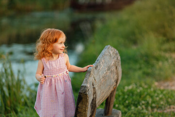 Cheerful red-haired toddler girl in a pink dress standing barefoot on a large rustic log bench,...