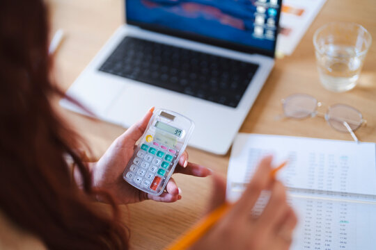 An Arab woman reviews financial data. She uses a calculator and pencil to analyze the numbers on a spreadsheet. A laptop and glasses are on the desk.