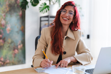Smiling Arab businesswoman works at her desk. She is reviewing documents and using a pencil to make notes, with a laptop nearby.