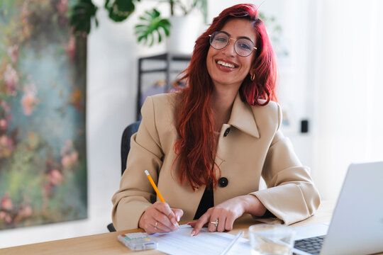 Smiling Arab businesswoman works at her desk. She is reviewing documents and using a pencil to make notes, with a laptop nearby.