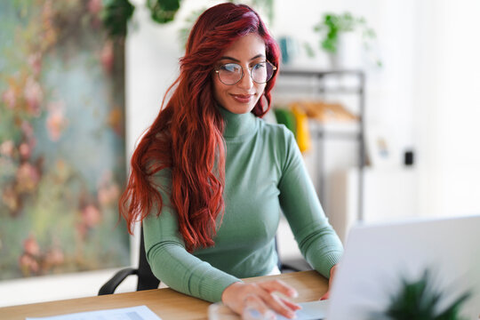 An Arab woman works on her laptop at a desk. She is wearing glasses and a green turtleneck, focused on her work.