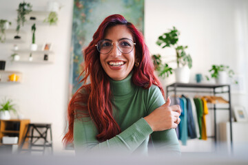 An Arab woman with red hair smiles warmly at the camera. She is wearing glasses and a green turtleneck sweater in a bright, modern home.