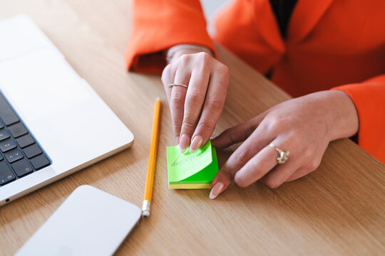 An Arab woman is working at her desk. She is writing notes on a sticky note next to her laptop and phone, wearing an orange blazer.