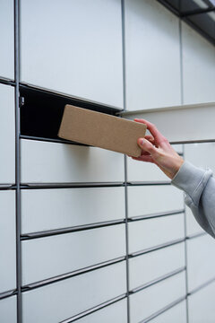 Young man placing a cardboard parcel into a self-service parcel locker outdoors. Close-up of a person inserting a package into an open locker of an outdoor parcel delivery station