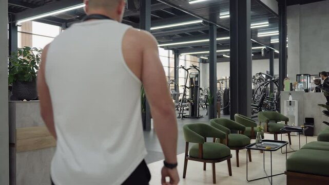 Well bodied man walks into modern gym through reception hall as people exercise on equipment, weights, cardio machines and free training areas under bright natural light from large glass windows