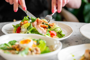 A close-up of a fresh salad with lettuce, tomatoes, cucumbers, bacon, and a soft-boiled egg on a white plate. A person uses a knife and fork to eat, emphasizing healthy eating and vibrant colors.