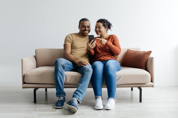 Obraz premium Beautiful Young Black Couple Using Mobile Phone Together At Home, Happy African American Spouses Relaxing On Couch With Modern Smartphone, Browsing New App Or Shopping Online, Copy Space