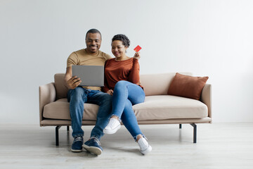 Obraz premium Online Shopping Concept. Happy Black Couple Using Credit Card And Laptop While Sitting On Couch Over White Background In Studio, Cheerful Man And Woman Enjoying Purchasing In Internet
