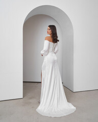 Brunette woman in wedding dress poses elegantly in bright interior with arched backdrop