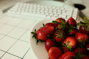 Close view shot of a white computer keyboard next to a bowl of ripe, glossy strawberries on a grid-patterned desk, blending technology with a fresh, healthy snack.