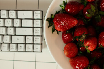Overhead close-up of a white bowl brimming with ripe red strawberries beside a compact white...
