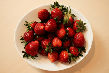 A top-down view of a white bowl filled with fresh, vibrant red strawberries on a light wood surface, highlighting their glossy texture and bright green calyxes in a simple, appetizing composition.