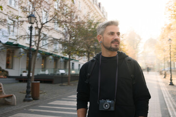 Obraz premium Man exploring a picturesque sunny city street with camera and thoughtful expression.