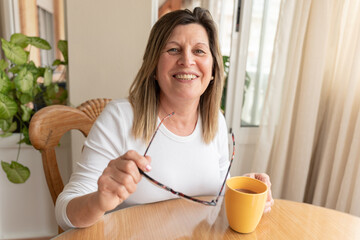 Smiling middle-aged woman wearing reading glasses, sipping coffee while seated at wooden dining...