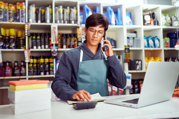 Salesman wearing apron taking orders by phone and calculating total price in auto parts store