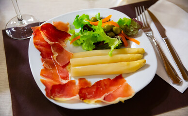 Slices of jamon served with asparagus, lettuce and carrot on white plate in restaurant.