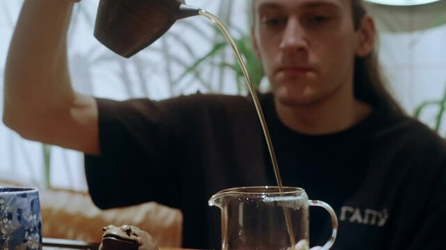 A tea master carefully pours brewed tea from a clay teapot into a glass teapot during a traditional ceremony on a wooden tray.