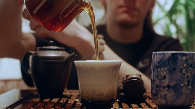 A tea master gracefully pours brewed tea from a glass teapot into two ceramic bowls during a traditional tea ceremony.