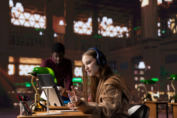 Young girl listening to an educational documentary on her laptop and taking notes, using modern non conventional way to study in the quietness of university library. Student collects academic data.