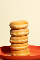 Five perfectly stacked glazed donuts rest on a vivid orange plate against a neutral beige background, capturing a clean, balanced, and visually tempting moment of sweet minimalism.