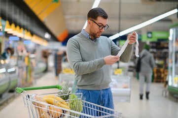 Shocked caucasian man with rounded eyes looking at extremely long bill. Amazed buyer doing grocery...