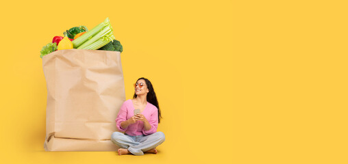 A woman sits on the floor beside a large paper bag filled with colorful vegetables and fruits,...