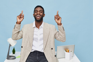 Happy joyful man with dark skin wearing beige blazer and white shirt, raising both hands with fingers pointing up, standing in modern office with desk, lamp, and supplies on blue background. People
