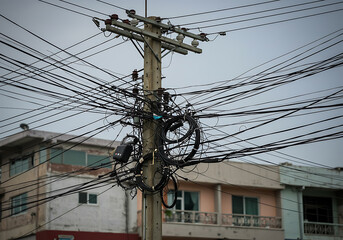 An overabundance of wires clutters a concrete utility pole in an urban setting, with a cloudy sky.