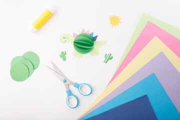 Handmade paper dinosaur and craft supplies on a white background. Flat lay composition with scissors, glue stick, colored paper, and cut-out shapes. DIY children's craft and creative hobby concept,
