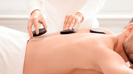 Fototapeta premium Tranquil middle aged muscular man having hot stone massage at modern spa, side view. Spa female therapist placing black stones on relaxed businessman back, healing massage at spa salon