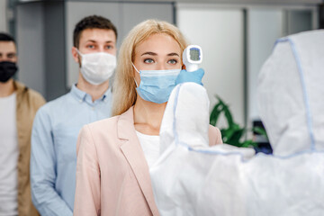 Health control at work and new normal for company. Man in protective suit checks temperature with...