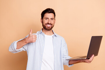 Confident young man with beard in stylish attire holding laptop and showing thumbs-up gesture against beige background