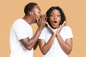 African-american guy sharing secret or whispering gossips into his girlfriend's ear, yellow studio background. Black man whispering words of love to his amazed woman who screaming and touching cheeks