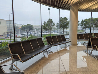 Modern empty airport chairs seating area interior with wide glass windows and rainy day exterior view with parked buses and urban scenery. Contemporary architecture, travel waiting, quiet space. 