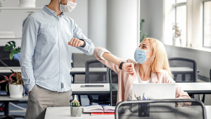 Modern greeting at office during coronavirus epidemic and protection from disease. Young man and woman in protective masks touch with elbows to say hello in interior of office, empty space