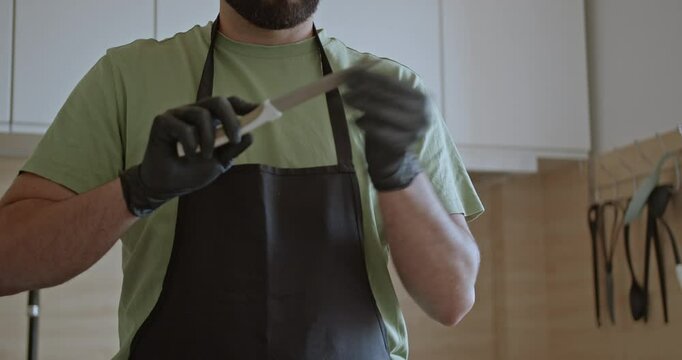 A man in an apron and black gloves sharpens a knife in his home kitchen. He tests the sharpness with his finger.