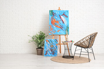 Armchair, table with art supplies and paintings near white brick wall in room