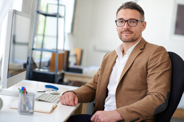 Obraz premium Portrait, glasses and man on desk, keyboard and vision for project, reporter and professional. Face, proud and entrepreneur in company, journalist and confidence in business and notes for research