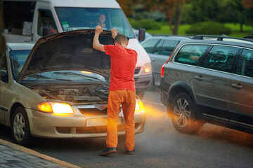 Man opens hood of smoking car on roadside after engine overheats due to coolant leak, surrounded by passing cars. Man stands beside broken down car with raised hood in busy city traffic in the evening
