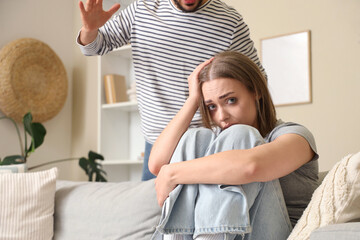 Crying young woman sitting on sofa and her husband at home. Domestic violence concept