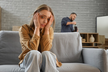 Scared young woman sitting on sofa and her drunk husband at home. Domestic violence concept