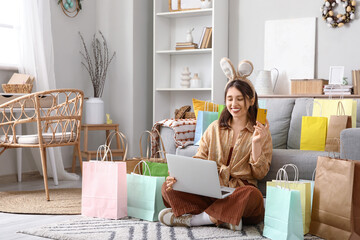 Young woman in bunny ears with credit card and laptop shopping online at home on Easter Day