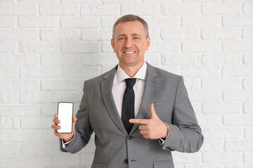 Portrait of mature businessman pointing at modern mobile phone with blank screen near white brick...
