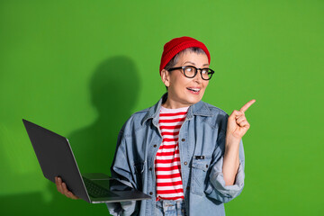 Stylish older woman in casual denim and beanie holding a laptop in front of a green background,...